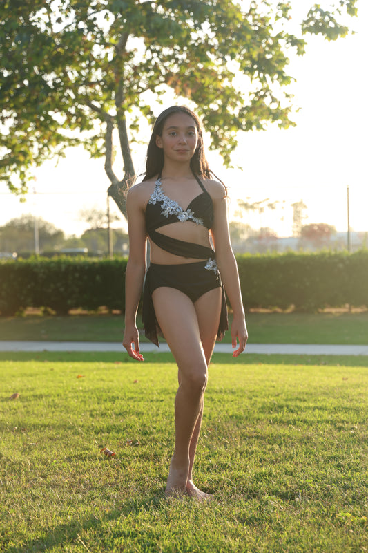 Dancer in a black and silver dance costume standing on grass with trees and water in the background